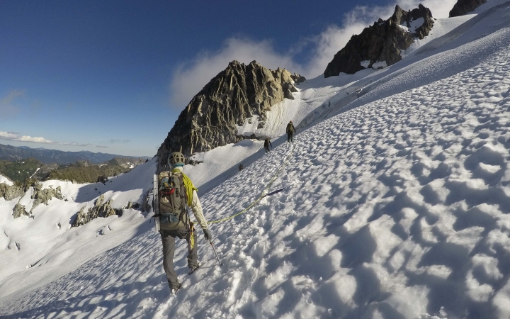 A person wearing a backpack and snow gear, hikes across a snow incline. There are others in front of them. They appear to be at high elevation.